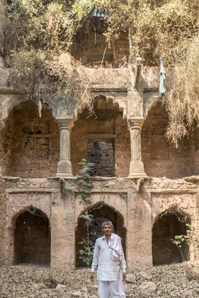Man standing in front of an ancient architectural structure with arched niches and overgrown vegetation.