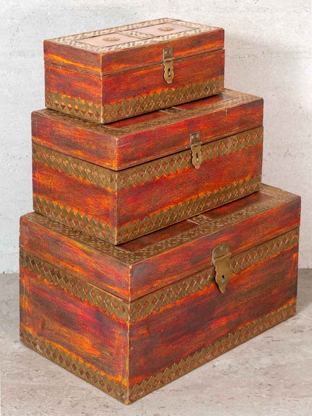 Three stacked wooden storage chests in different sizes, decorated with a distressed red and orange wood finish and metallic gold geometric borders, set against a neutral concrete background.