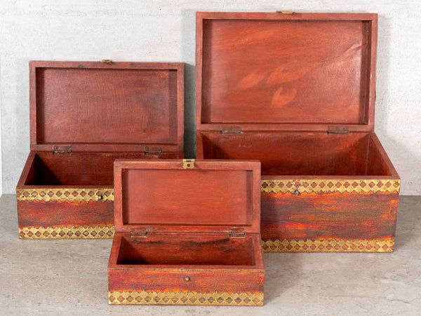 Three vintage wooden storage boxes of different sizes, all open to show their interiors, decorated with gold and black geometric patterns along the lower front edges, set against a light gray concrete background.