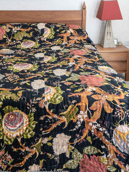 A black quilted bedspread with an intricate, colorful botanical and bird pattern, styled on a bed with a wooden headboard and bedside lamp.