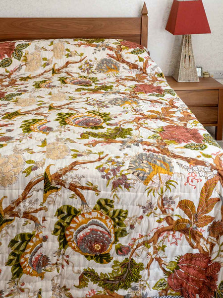 A quilted bedspread with an intricate botanical and bird pattern in earthy tones of green, brown, rust red, and beige, styled on a bed in a bedroom setting.