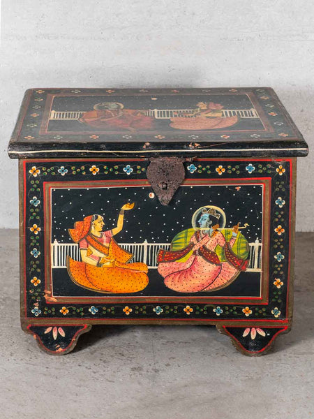 A vintage Indian wooden chest decorated with hand-painted Madras style scenes of Lord Krishna and Radha under a starry night sky, on a gray concrete floor.