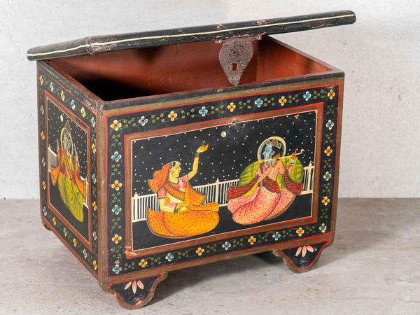 An antique Indian wooden storage box with a hinged lid, featuring hand-painted folk art illustrations of Lord Krishna and Radha under a starry night sky on a black background.