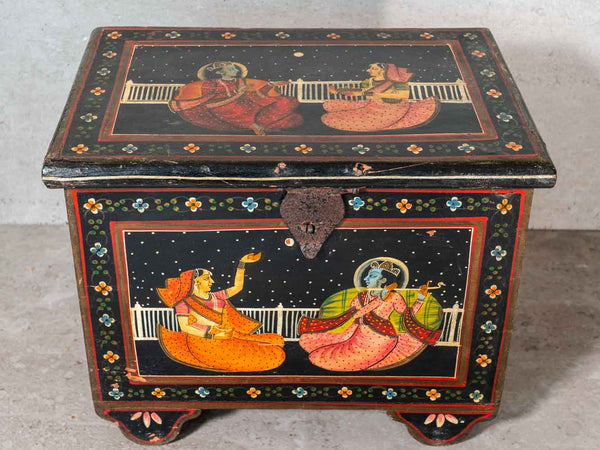 A traditional Indian hand-painted wooden chest with a black lacquered finish, featuring colorful illustrations of deities in orange and pink garments on a starry night sky background, set against a neutral concrete wall.