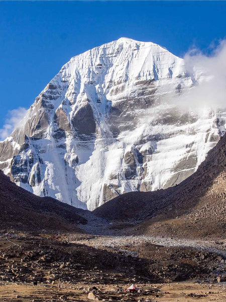 A majestic snow-covered mountain peak rises under a clear blue sky, with rocky brown slopes in the foreground and a small structure near the bottom center.
