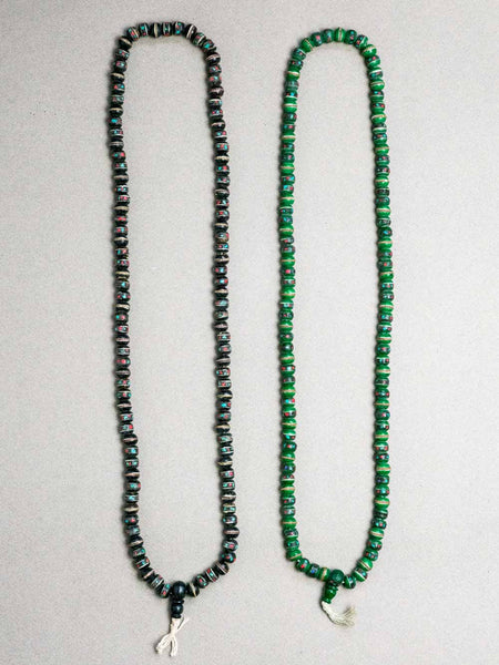 Two beaded Tibetan malas on a gray background. The left necklace has dark beads with a subtle iridescent sheen, and the right necklace has bright green beads with a textured, marbled pattern.