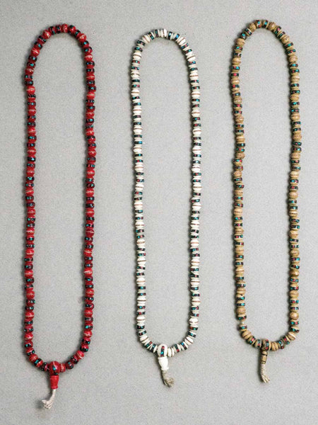 Three long beaded prayer beads on a light gray background. The left necklace is red and black, the middle is white and black, and the right is tan and black.