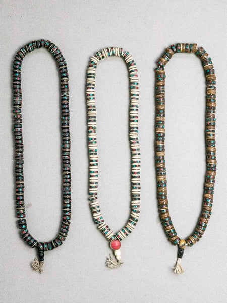 Three beaded Tibetan malas. The left mala is black with turquoise and red accents, the middle is white with turquoise and red accents and a small pink bead, and the right is brown and gold with turquoise and red accents.