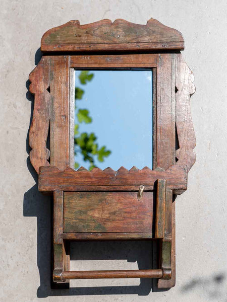 A rustic, hand-carved wooden wall mirror cabinet with a small lock and shelf, mounted on a textured light gray wall.