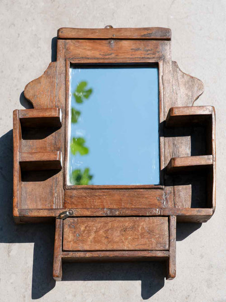 A rustic wooden wall-mounted mirror cabinet with side shelves and a single drawer, photographed against a light concrete wall with reflections visible in the mirror.